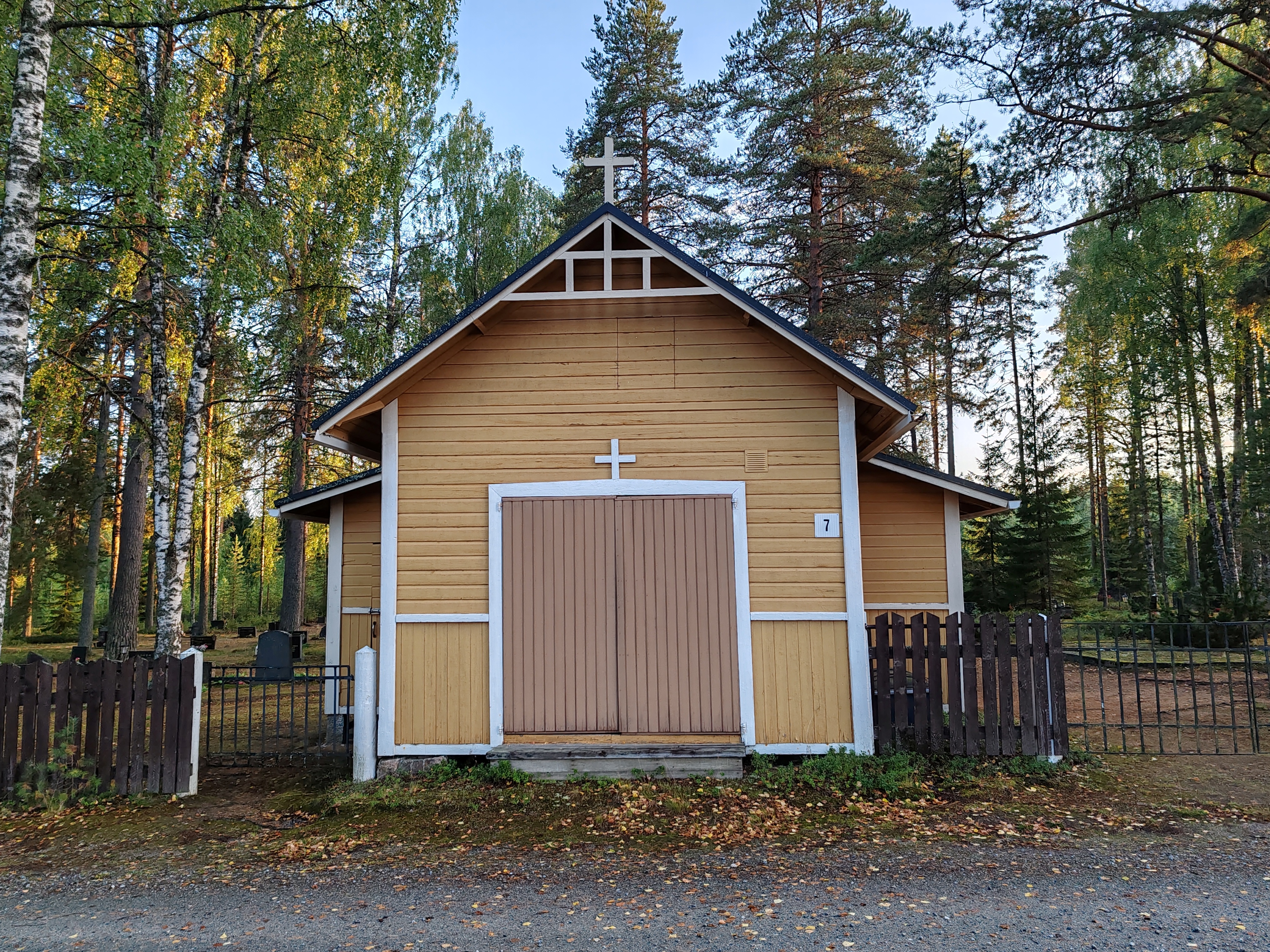 Höljäkän-Porosaaren hautausmaa Kuvassa puinen rakennus, jonka harjalla risti. Rautaportit molemmilla puolilla ja taustalla häämöttää hautamuistomerkkejä.