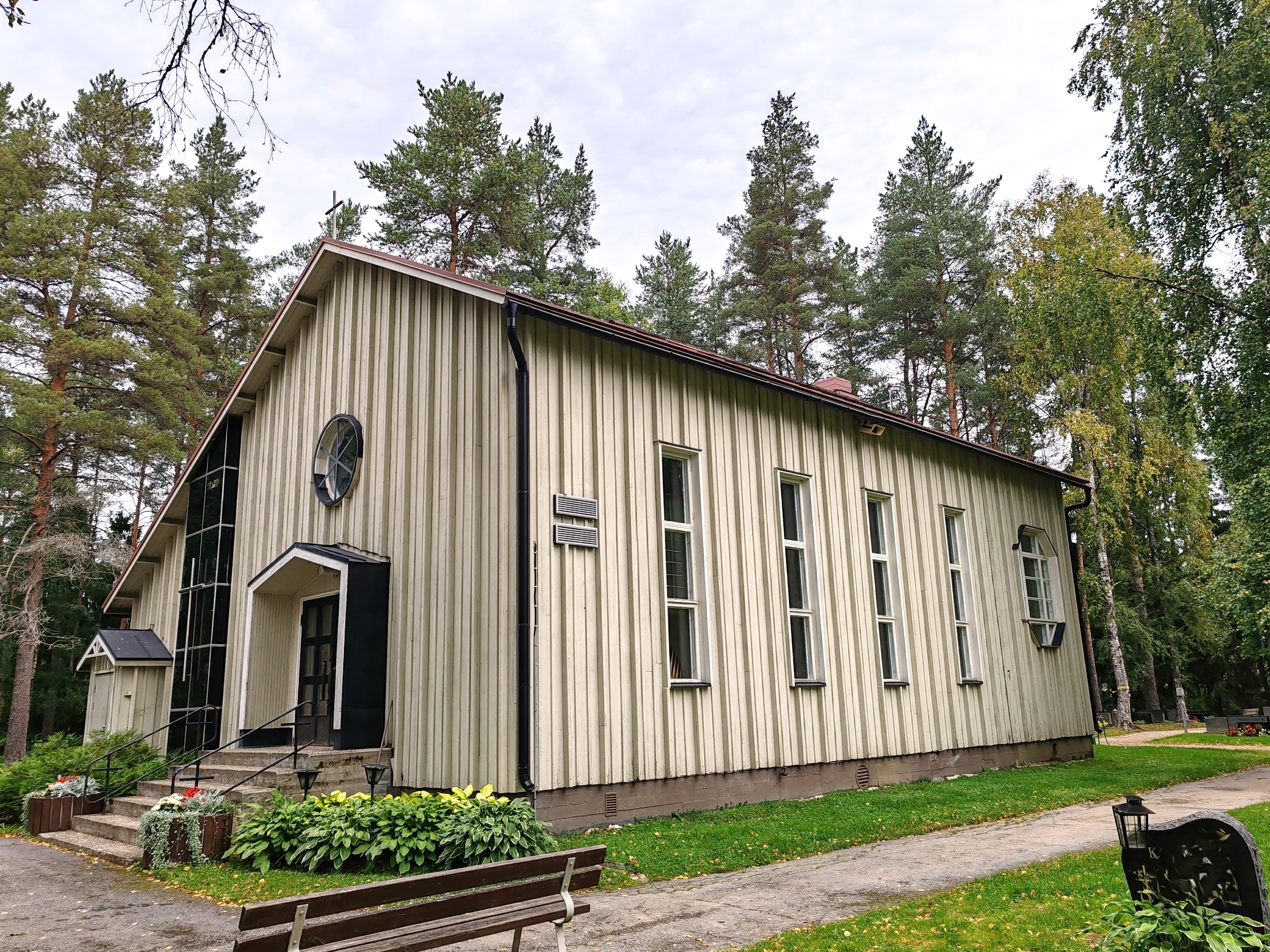 Valtimon siunauskappeli Kuvassa puinen rakennus, ympärillä kukkaistuksia ja hautakivi ja penkki, Valtimon siunauskappeli.